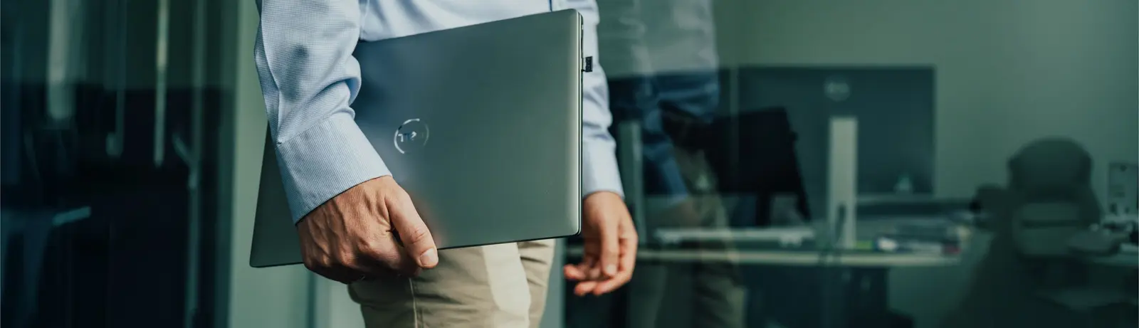 Person hält einen geschlossenen Laptop an der Seite in einem Büro mit Schreibtischen im Hintergrund.