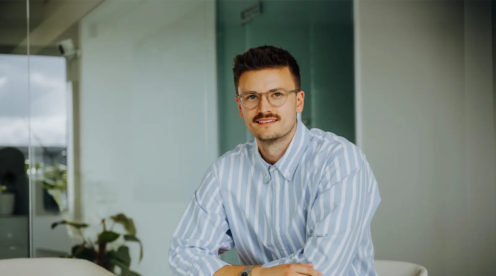 Junger Mann mit Brille und gestreiftem Hemd sitzt lächelnd in einem modernen Büro.