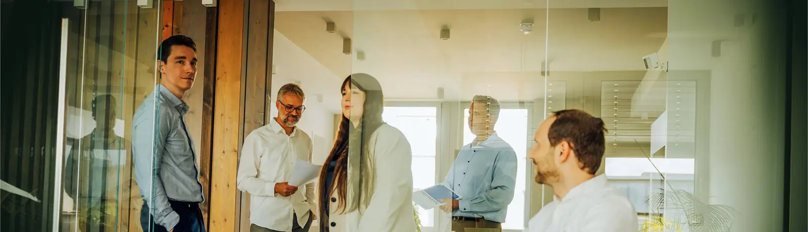 Fünf Personen im Büro, die sich unterhalten und Unterlagen halten, durch eine Glaswand gesehen.