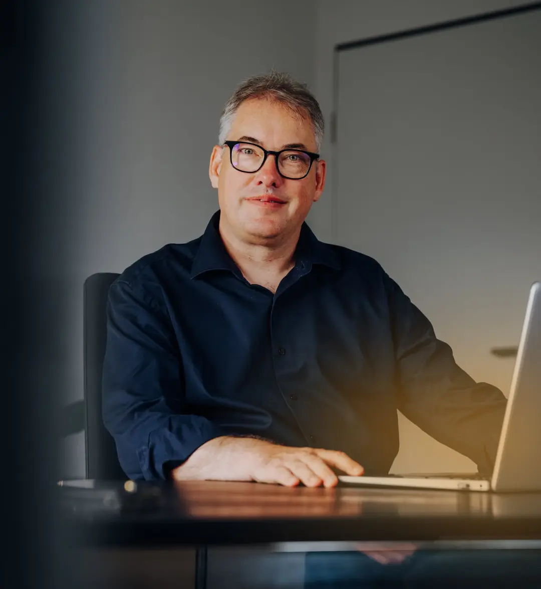 Mann mit Brille und blauem Hemd sitzt an einem Tisch mit Laptop und lächelt in die Kamera.