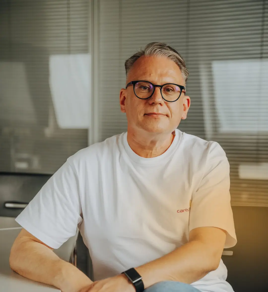 Mann mit Brille und weißem T-Shirt sitzt entspannt an einem Tisch vor Fenster mit Jalousien.