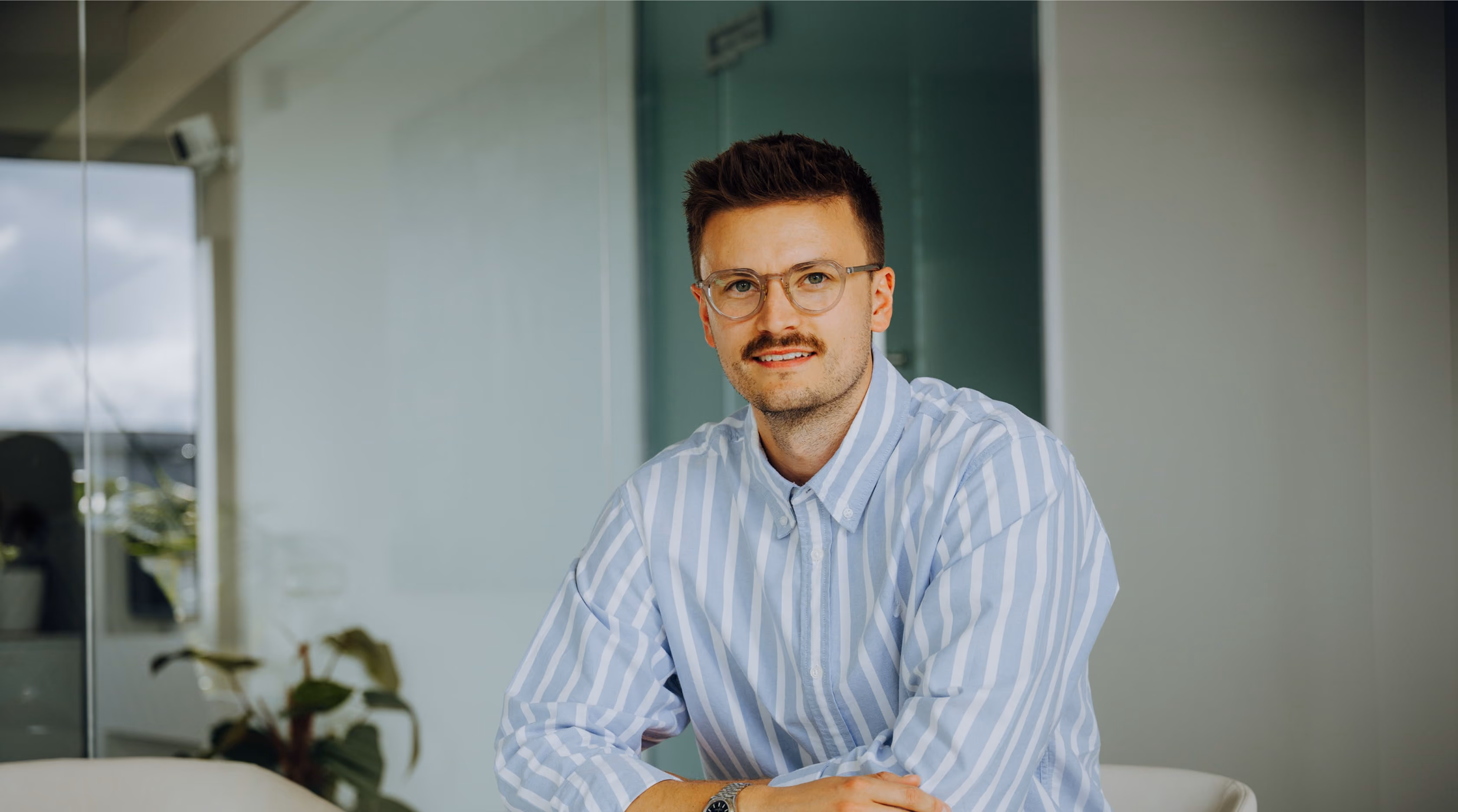 Junger Mann mit Brille und gestreiftem Hemd sitzt lächelnd in einem modernen Büro.