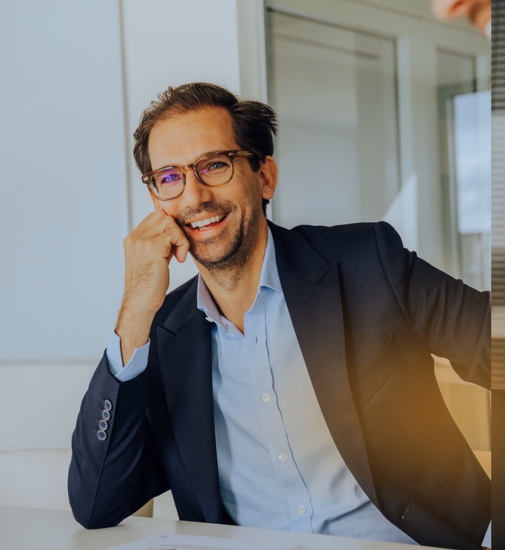 Lächelnder Mann mit Brille und Anzug lehnt sich an den Tisch in einem modernen Büro.