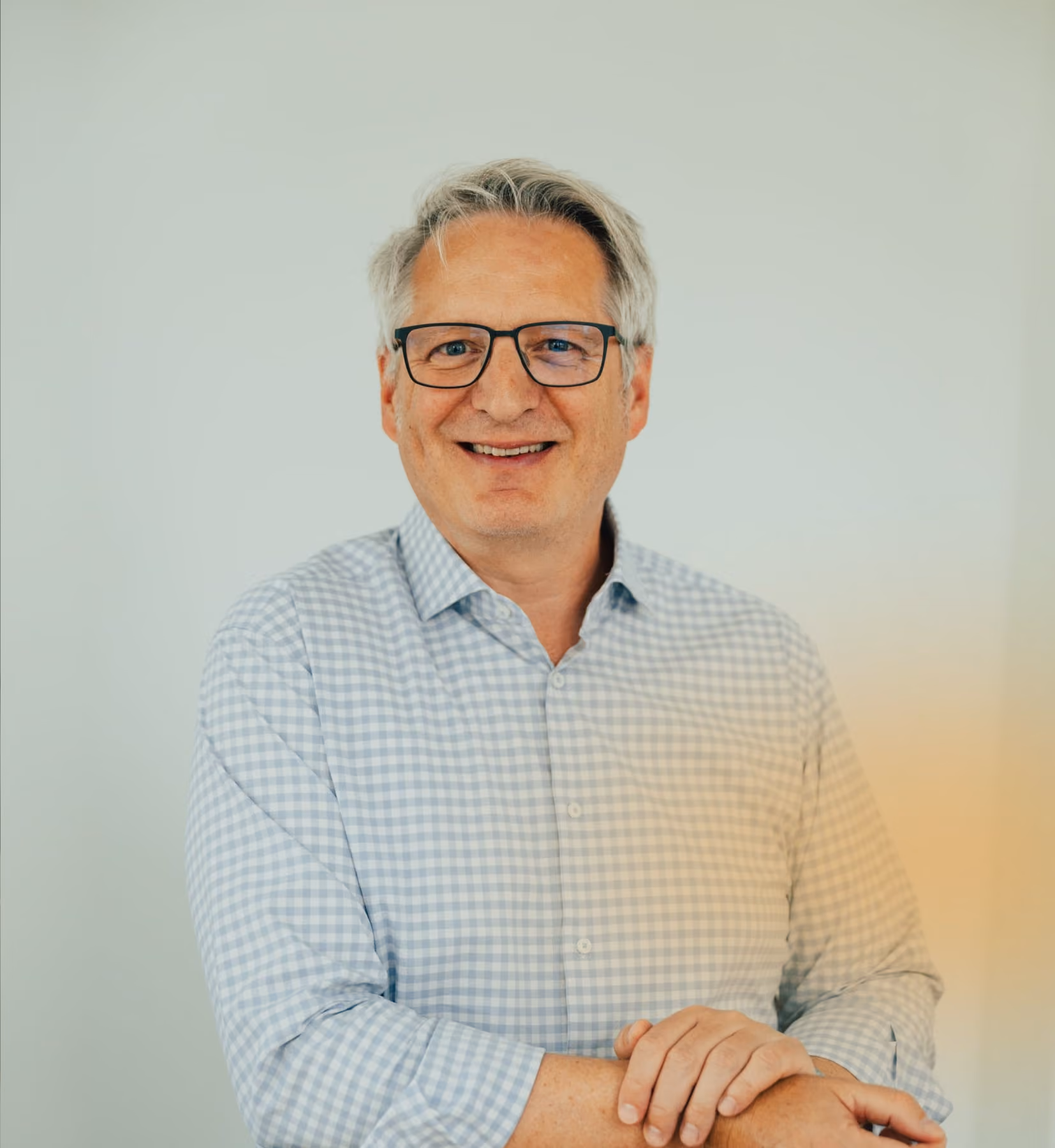 Porträt eines lächelnden älteren Mannes mit Brille und kariertem Hemd vor hellem Hintergrund.