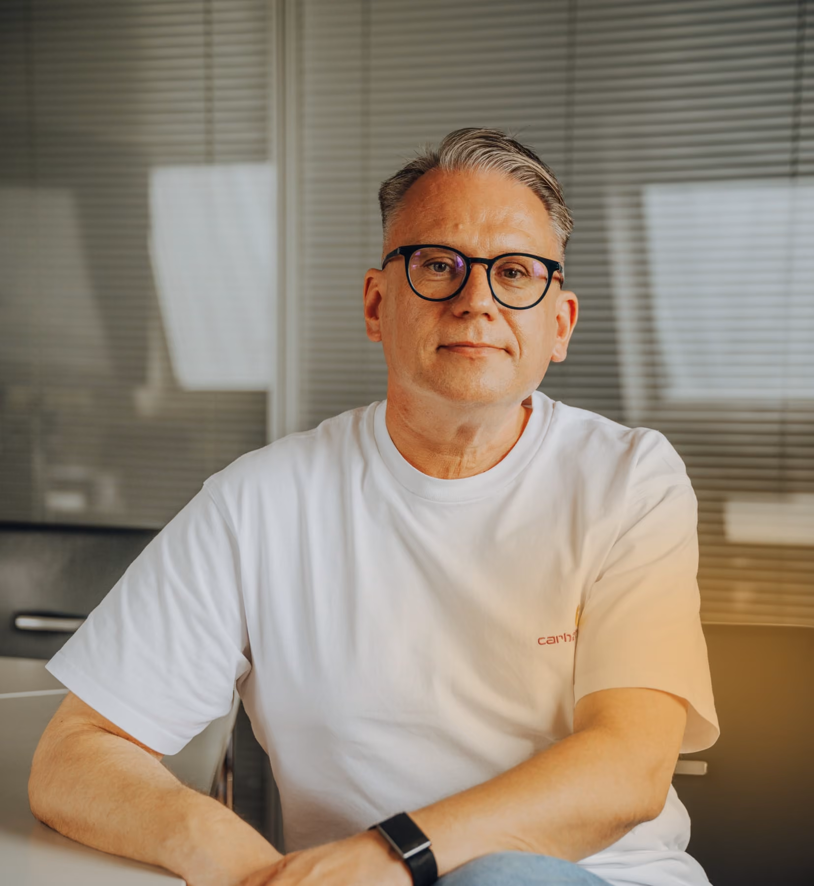 Mann mit Brille und weißem T-Shirt sitzt entspannt an einem Tisch vor Fenster mit Jalousien.