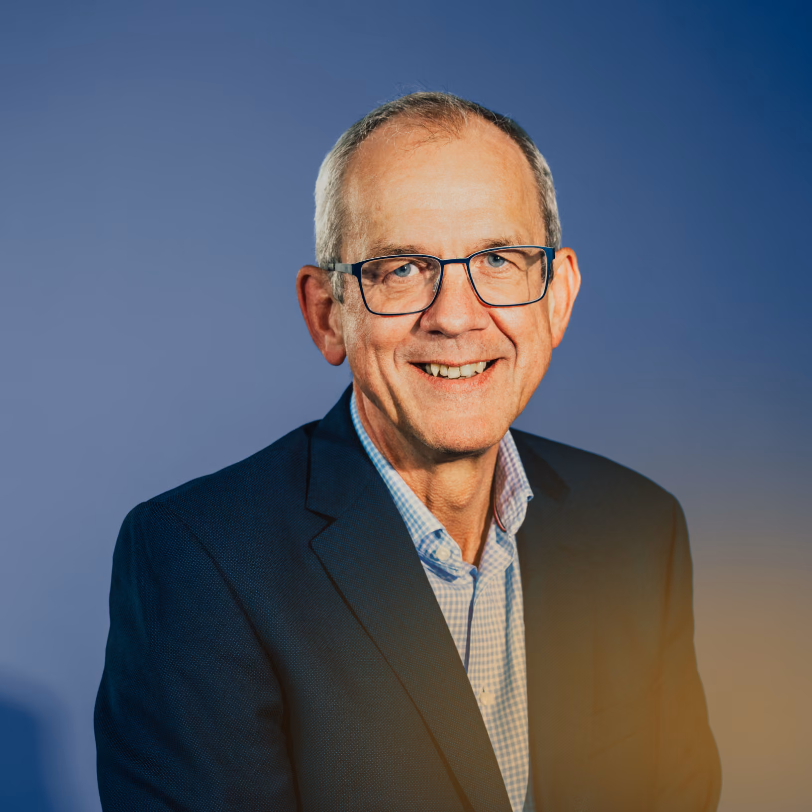 Älterer Mann mit Brille, lächelnd, trägt blauen Anzug und kariertes Hemd vor blauem Hintergrund.