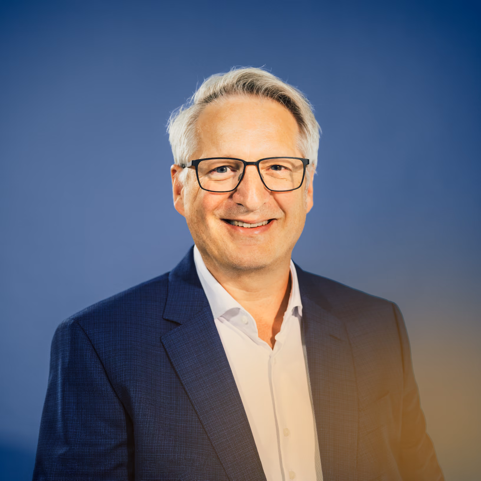 Älterer Mann mit Brille, der in einem dunkelblauen Anzug und weißem Hemd vor blauem Hintergrund lächelt.
