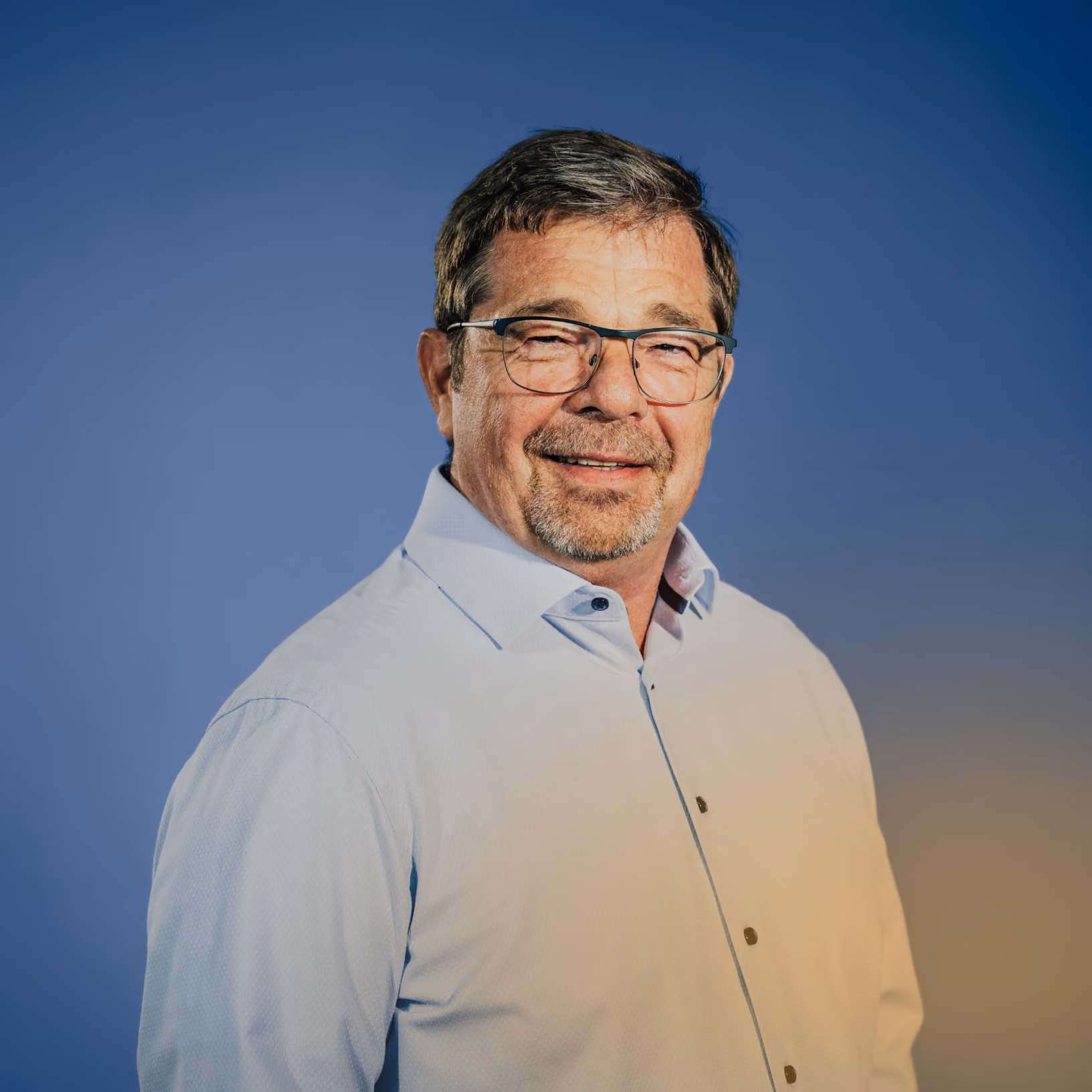Älterer Mann mit Brille, grauem Haar und Bart, lächelnd in weißem Hemd vor blauem Hintergrund.