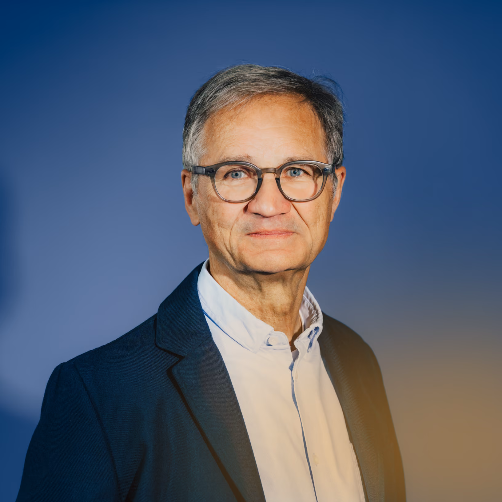 Porträt eines älteren Mannes mit grauem Haar, Brille, weißem Hemd und dunkelblauem Sakko vor blauem Hintergrund.