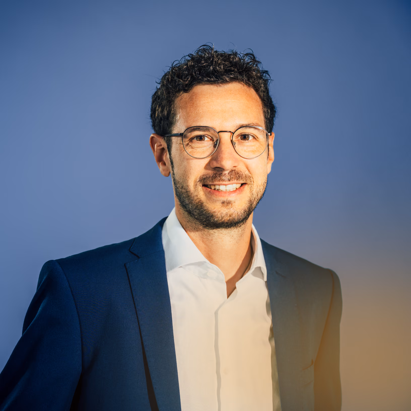 Mann mit kurzer dunkler lockiger Haare, Brille, blauem Sakko und weißem Hemd lächelt vor blauem Hintergrund.