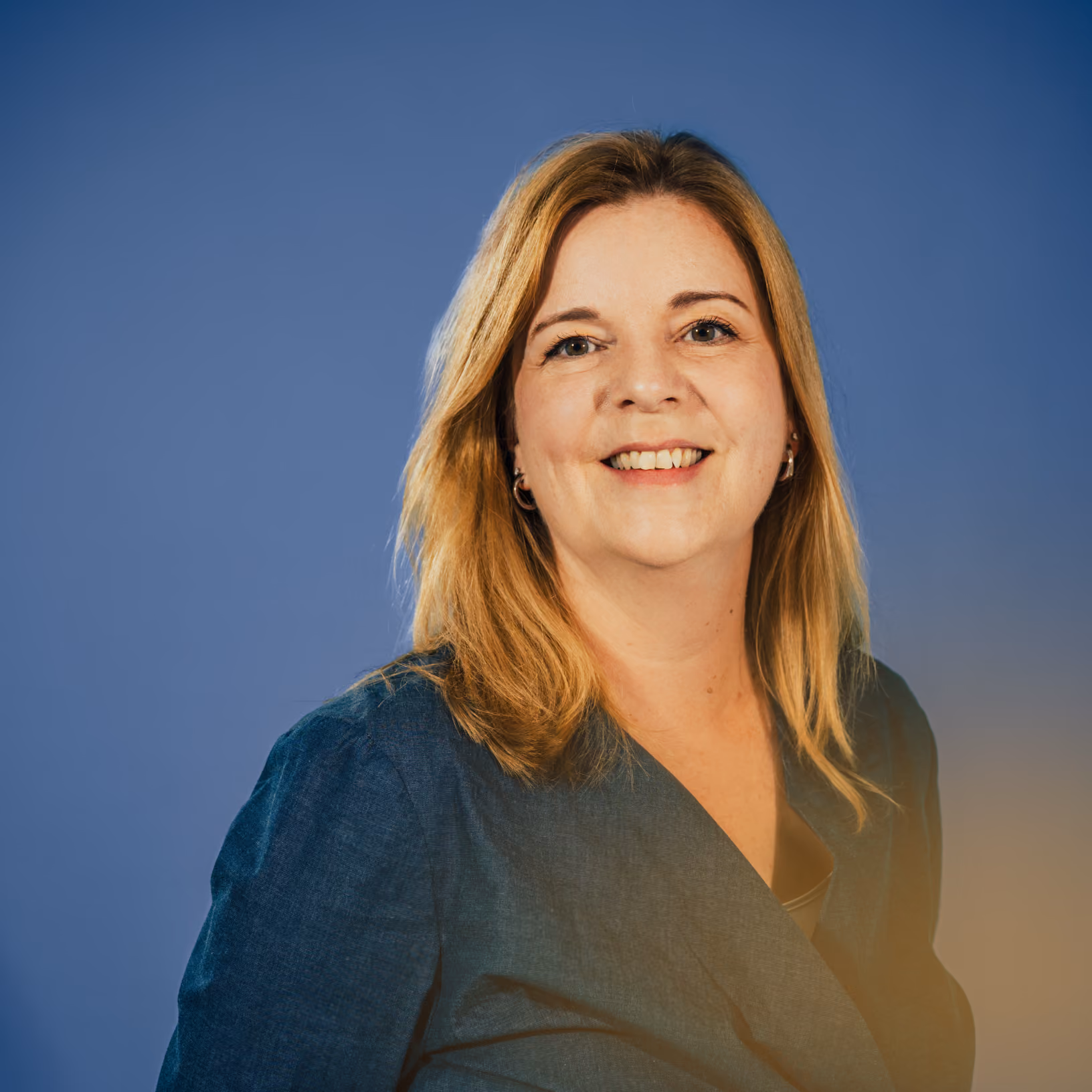 Lächelnde blonde Frau in dunkelblauer Bluse vor blauem Hintergrund.