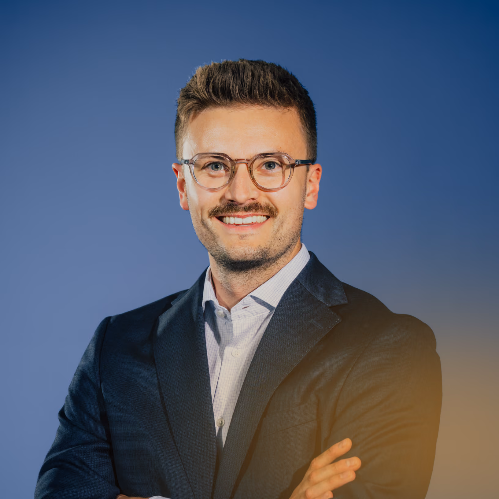 Mann mit Brille, Anzug und Hemd lächelt mit verschränkten Armen vor blauem Hintergrund.