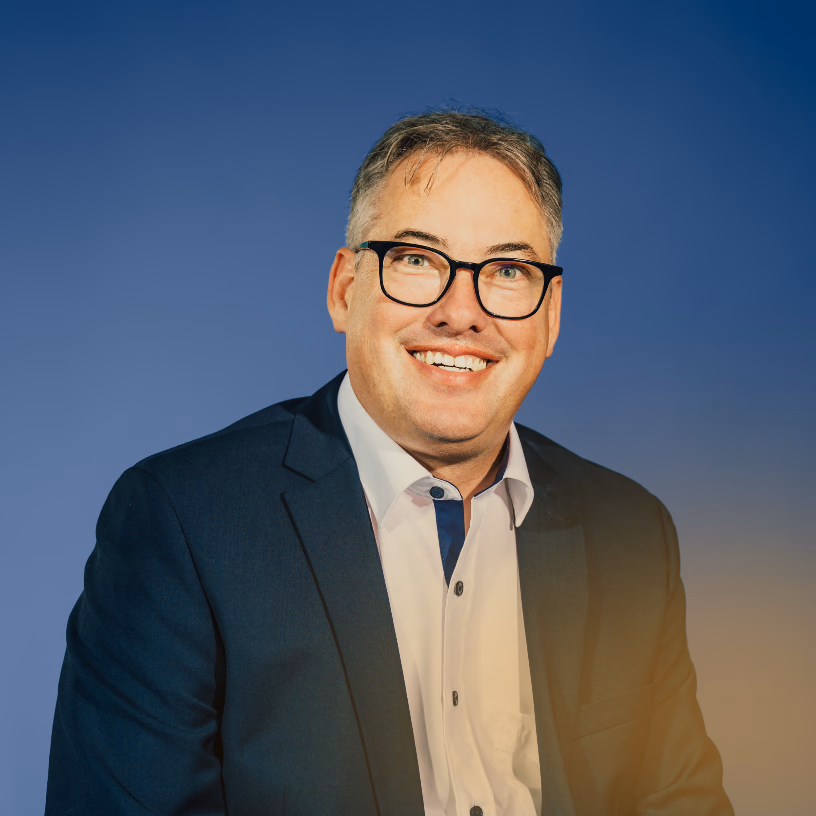 Porträt eines lächelnden Mannes mit Brille, dunklem Anzug und weißem Hemd vor blauem Hintergrund.