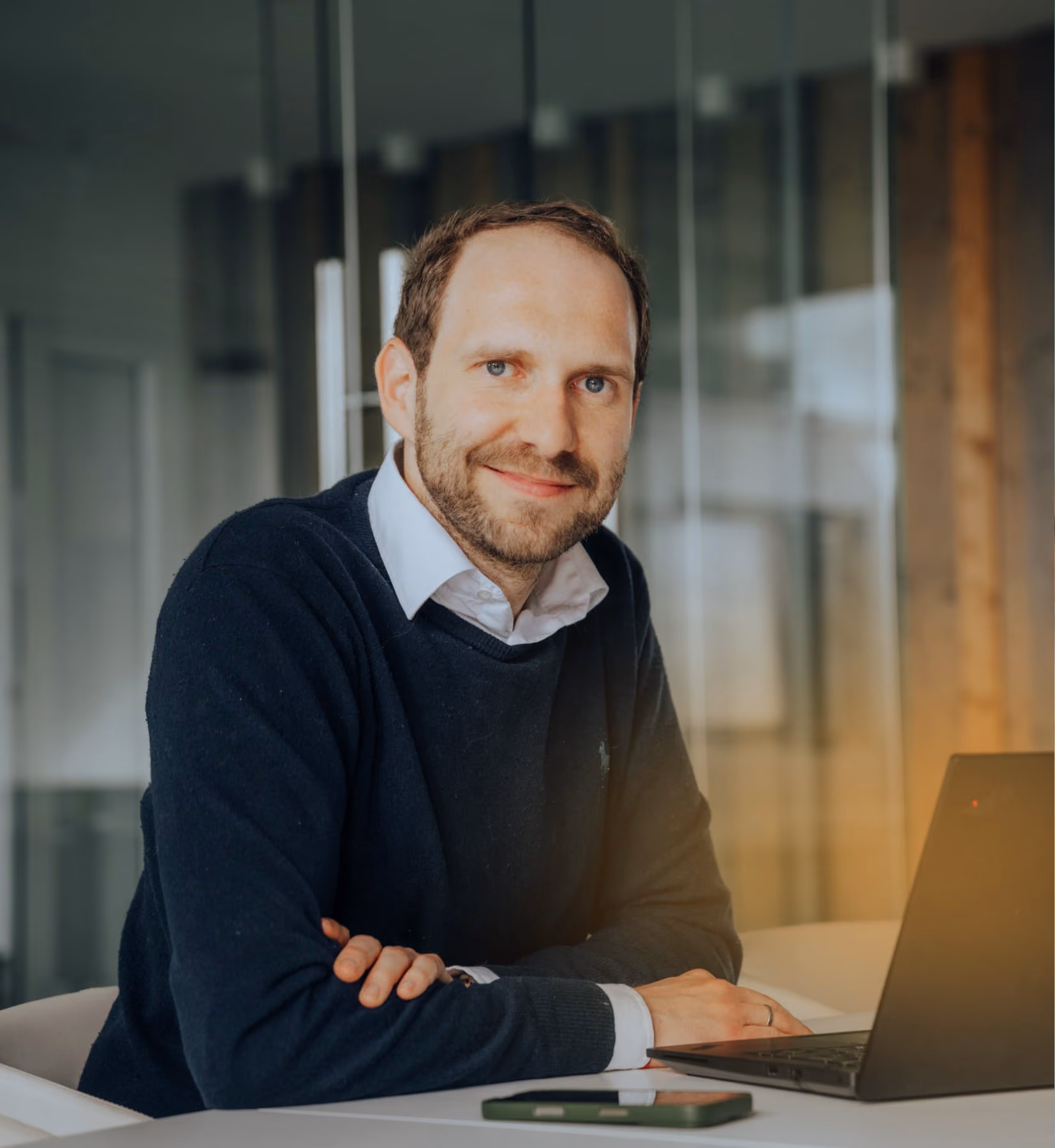 Mann mit blauem Pullover sitzt lächelnd am Tisch vor Laptop und Smartphone im Büro.
