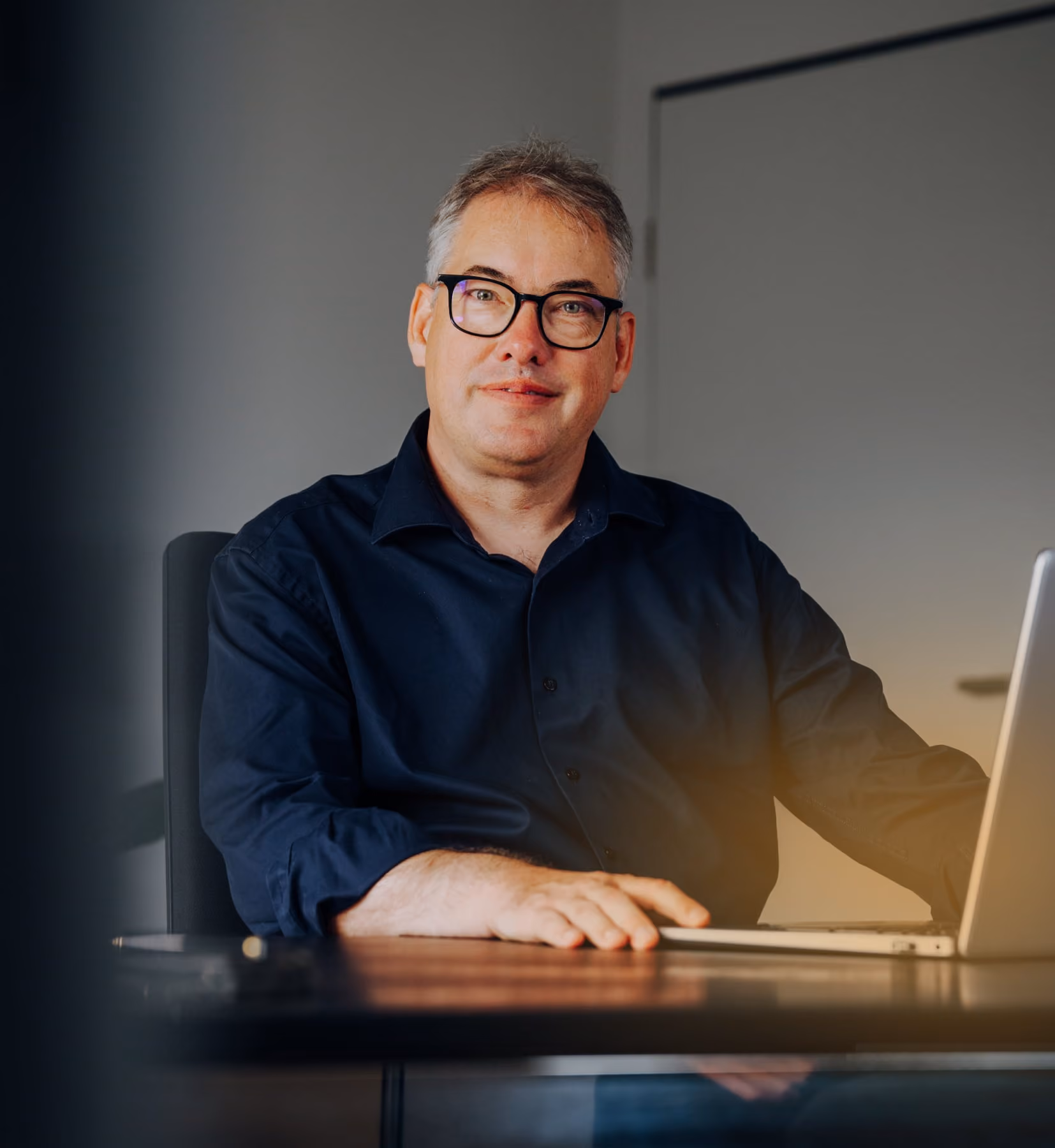 Mann mit Brille und blauem Hemd sitzt an einem Tisch mit Laptop und lächelt in die Kamera.