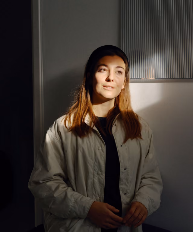 Junge Frau mit braunen Haaren im hellen Mantel steht an Wand mit Licht und Schatten.