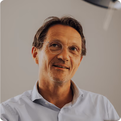 Porträt eines lächelnden Mannes mittleren Alters mit Brille und weißem Hemd vor grauem Hintergrund.